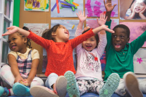 Four young children sit on the floor and raise their hands with excitement